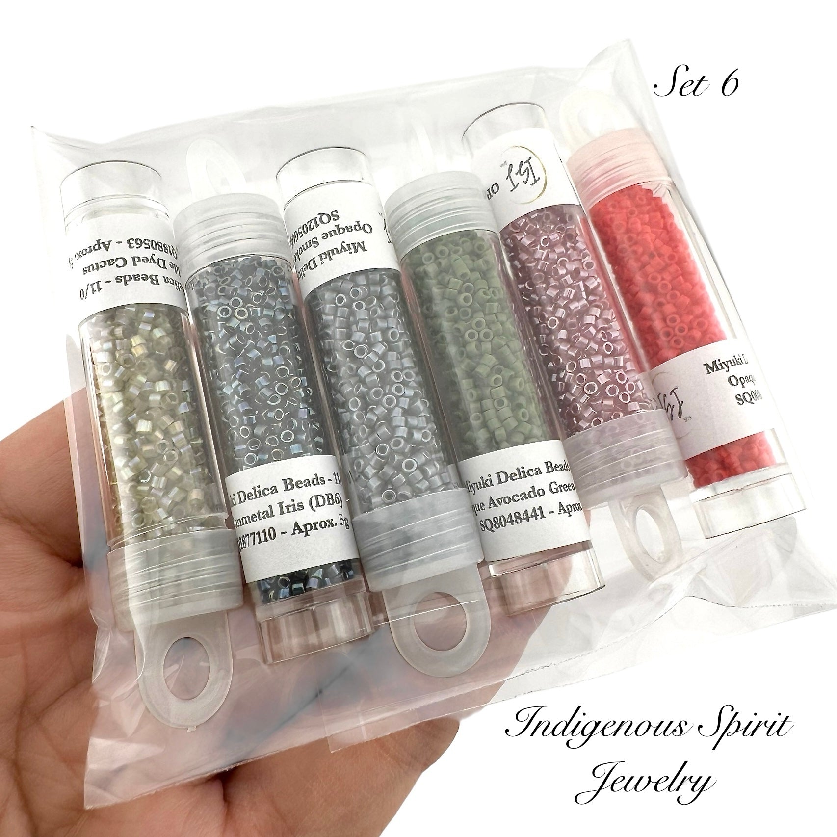 Set of six small cylindrical containers with beads, held in a hand against a white background. Packaging labeled 'Indigenous Spirit Jewelry'.