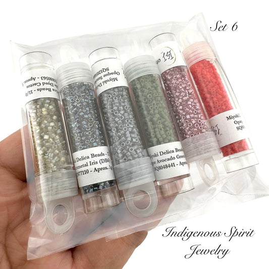 Set of six small cylindrical containers with beads, held in a hand against a white background. Packaging labeled 'Indigenous Spirit Jewelry'.
